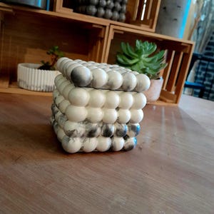May include: A square decorative box with a lid, featuring a unique design of rounded, bubble-like shapes. The box is primarily off-white with grey marbling, and sits on a wooden surface. The box is a decorative item.