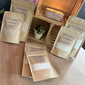 Peut inclure: Plusieurs petits sachets en papier brun remplis de poudre blanche sont présentés. Les sachets ont une fenêtre transparente et sont étiquetés en français. Une étagère en bois contient une bougie et une petite plante en pot. Thème: produits de parfum d'intérieur.