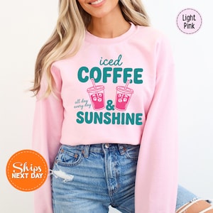 May include: Light pink sweatshirt with the words "iced COFFEE & SUNSHINE" in teal and white. Two iced coffee drinks are illustrated below the text. The phrase "all day every day" is above the coffee drinks. The sweatshirt is a crew neck.