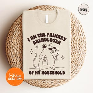 May include: An ivory t-shirt featuring a dark brown graphic of a mouse wearing sunglasses, holding a small bucket and a martini glass. The text reads, I AM THE PRIMARY BREADLOSER OF MY HOUSEHOLD, accompanied by small star icons surrounding the mouse.