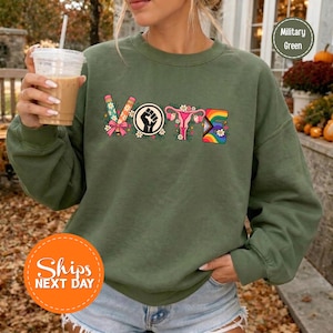 May include: A military green long-sleeve sweatshirt featuring the word VOTE in colorful graphic letters. The V is a pencil, the O is a black fist symbol, the T is a pink uterus, and the E is a rainbow-colored block.