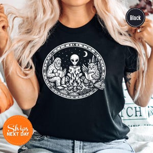 May include: Black t-shirt with a white graphic of a Bigfoot, an alien, and an owl reading books around a campfire under a starry night sky. The design is circular and includes a crescent moon and stars.