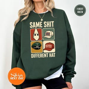 May include: Forest green sweatshirt with the text "SAME SHIT DIFFERENT HAT" and four images of different hats. The sweatshirt has a crew neck and long sleeves. The image includes a "Ships Next Day" sticker.