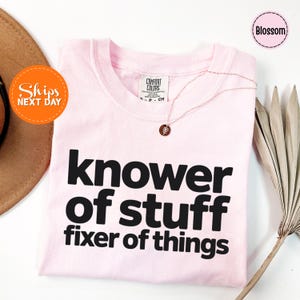 May include: A light pink t-shirt with the words "knower of stuff fixer of things" in bold black letters. A delicate necklace with a small pendant rests on the shirt. The shirt is folded and displayed on a white surface.
