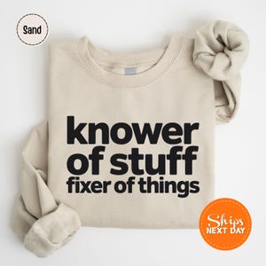 May include: A sand-colored sweatshirt with the text "knower of stuff fixer of things" in bold black letters. The sweatshirt is folded neatly, with a matching scrunchie resting on top. The words "Ships Next Day" are in an orange circle.