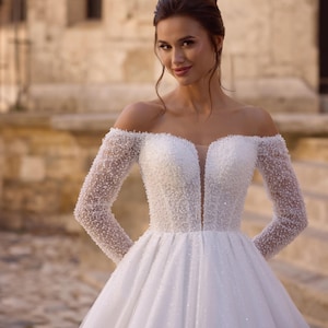 Op de afbeelding: Een witte off-the-shoulder trouwjurk met lange mouwen. De lijf en mouwen zijn versierd met kleine, glinsterende kralen. De rok is vol en vloeiend, met een subtiele glans. De jurk wordt gedragen door een persoon met donker haar, die buiten poseert.