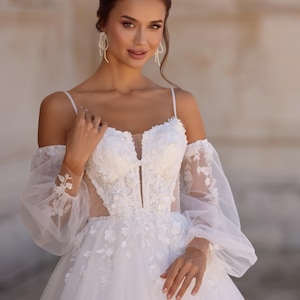 Op de afbeelding: Een witte trouwjurk met bloemenkanten details. De jurk heeft een sweetheart halslijn, doorschijnende off-the-shoulder mouwen en een volle rok. De jurk heeft een getailleerd lijfje en een zwierige rok. De afbeelding toont ook bungelende oorbellen.