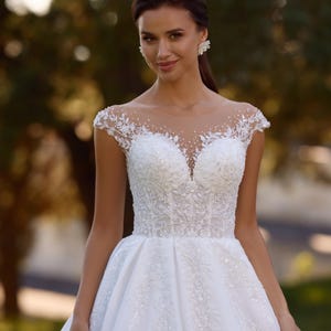 Op de afbeelding: Een witte trouwjurk met een doorschijnende halslijn en kapmouwen. Het lijfje is versierd met ingewikkeld kralenwerk en bloemenkant. De rok wijd uit vanaf de taille. De jurk heeft een klassiek, elegant ontwerp.