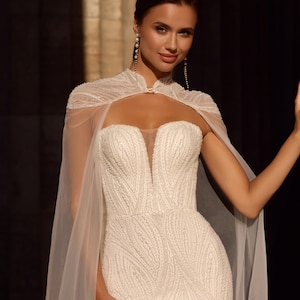 May include: A white, beaded wedding dress with a sheer cape. The gown has a sweetheart neckline and a fitted shape. The cape is attached at the shoulders. The image shows the details of the bridal gown. Long earrings complete the look. The dress is a classic design.