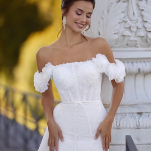 Op de afbeelding: Witte off-the-shoulder trouwjurk met bloemaccenten op de mouwen. De jurk heeft een getailleerd lijfje met sprankelende versieringen en een zwierige rok. Een dunne witte riem accentueert de taille. De afbeelding bevat het nummer "1057".