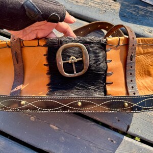 Könnte beinhalten: Handgefertigte Leder-Tragetasche mit einzigartigem Design. Die Tasche hat eine Kombination aus hellbraunem und schwarzem Leder, eine große Metallschnalle und dekorative Nähte. Die Tasche hat einen stabilen Griff und einen schwarzen Lederboden mit Metallnieten.