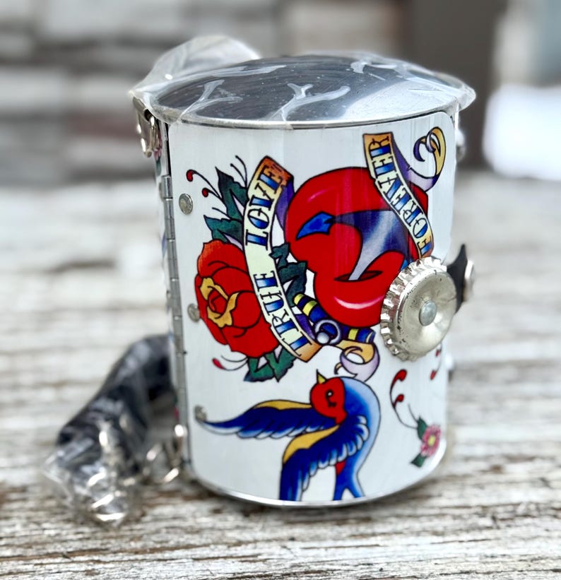 May include: A white metal container with a tattoo-style design featuring a red rose, a pair of red lips, and a blue swallow. The design includes the words "TRUE LOVE" and "FOREVER" on banners. The container has a silver lid and a silver bottle cap closure.
