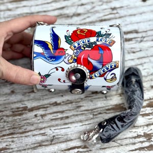 May include: A white, cylindrical purse with a tattoo-style design featuring a blue bird, red rose, and a heart. The text "TRUE LOVE FOREVER" is displayed. Includes a black strap and silver hardware.
