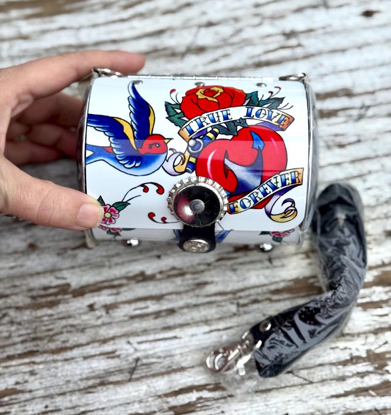 May include: A white, cylindrical purse with a tattoo-style design featuring a blue bird, red rose, and a heart. The text "TRUE LOVE FOREVER" is displayed. Includes a black strap and silver hardware.