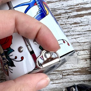 May include: A white, cylindrical container with a tattoo-style design featuring a blue swallow, red rose, and the word "LOVE". The container has a silver clasp and a black lanyard with a silver clip.