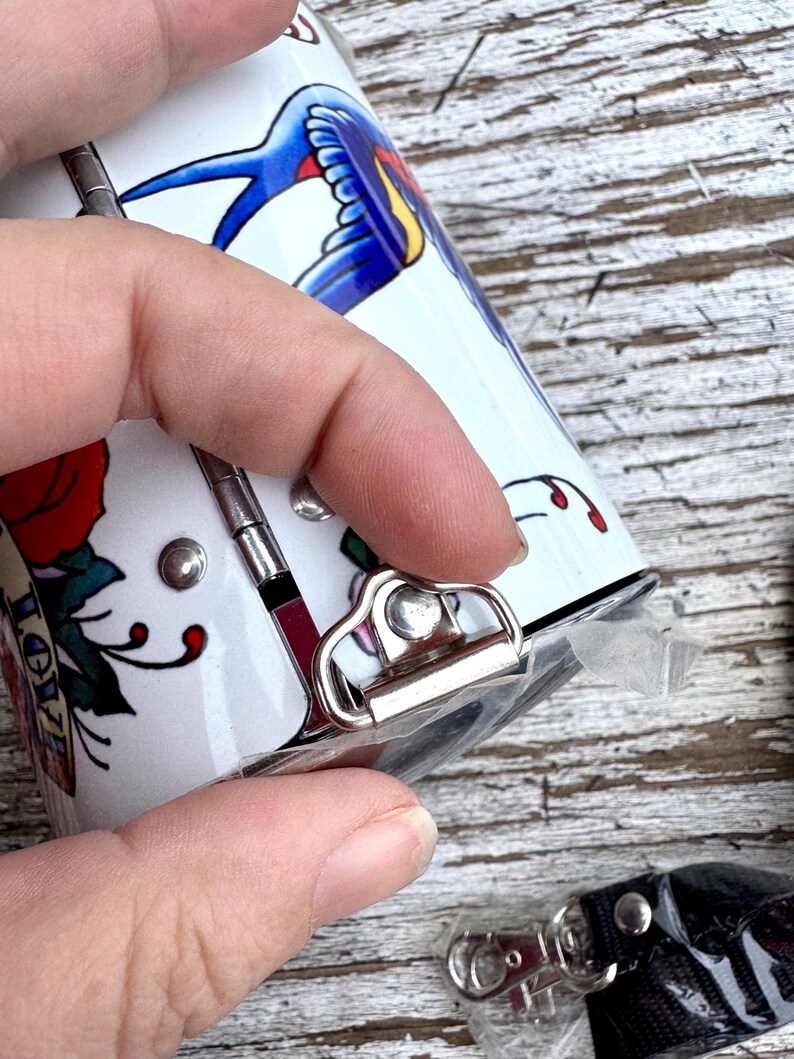 May include: A white, cylindrical container with a tattoo-style design featuring a blue swallow, red rose, and the word "LOVE". The container has a silver clasp and a black lanyard with a silver clip.