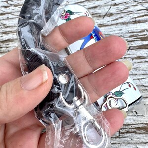May include: A black lanyard with a silver-colored clasp is partially visible, packaged in clear plastic. A white and floral-decorated object is in the background. The floral design includes pink and red flowers with green leaves.