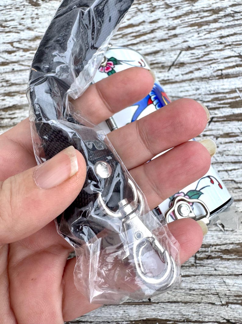 May include: A black lanyard with a silver-colored clasp is partially visible, packaged in clear plastic. A white and floral-decorated object is in the background. The floral design includes pink and red flowers with green leaves.