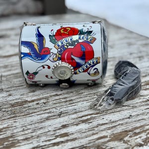 May include: A small, white, cylindrical purse with a tattoo-style design featuring a red rose, a blue swallow, and the words "TRUE LOVE FOREVER." The purse has a silver bottle cap closure and a black strap.