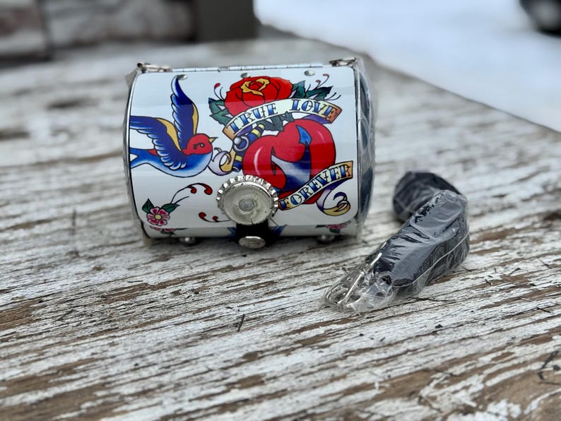 May include: A small, white, cylindrical purse with a tattoo-style design featuring a red rose, a blue swallow, and the words "TRUE LOVE FOREVER." The purse has a silver bottle cap closure and a black strap.