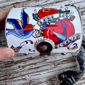 May include: White, cylindrical purse with a tattoo-style design. The design includes a blue bird, a red rose, and a heart with the words "True Love Forever" on banners. The purse has a silver-colored metal clasp and a black strap.