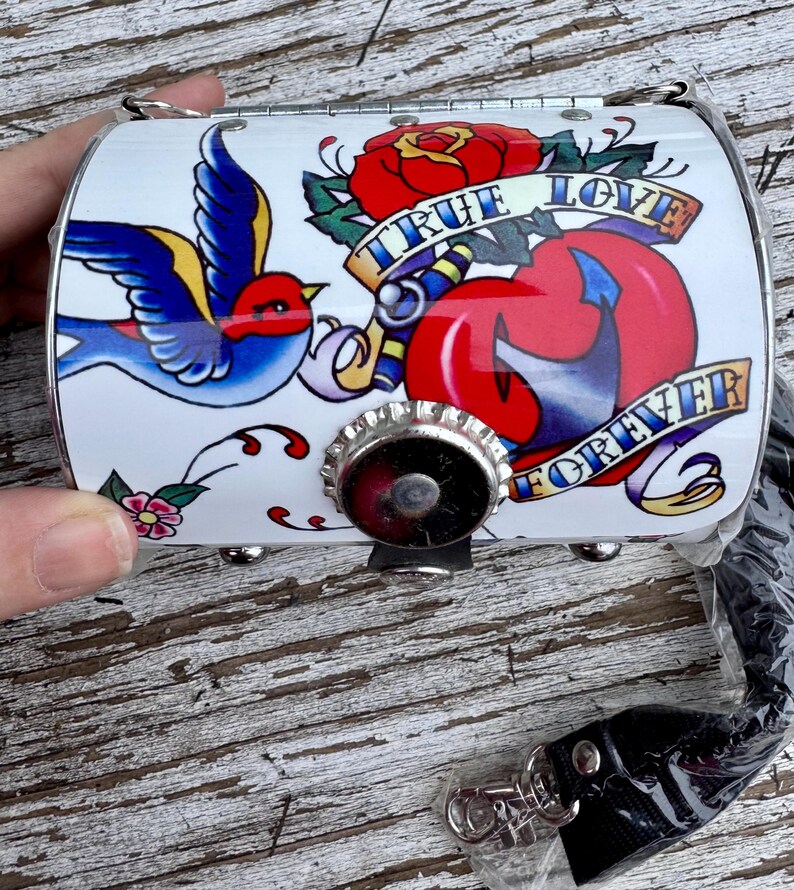 May include: White, cylindrical purse with a tattoo-style design. The design includes a blue bird, a red rose, and a heart with the words "True Love Forever" on banners. The purse has a silver-colored metal clasp and a black strap.
