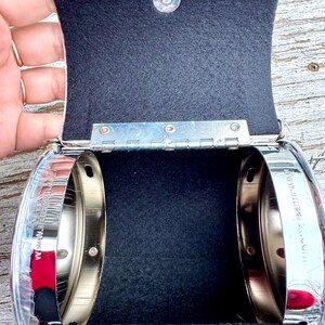 May include: Open, silver-toned metal purse with a black interior. The purse has a hinged lid with a magnetic closure. The bottom of the purse features a floral design with the text "WITH 6/07".