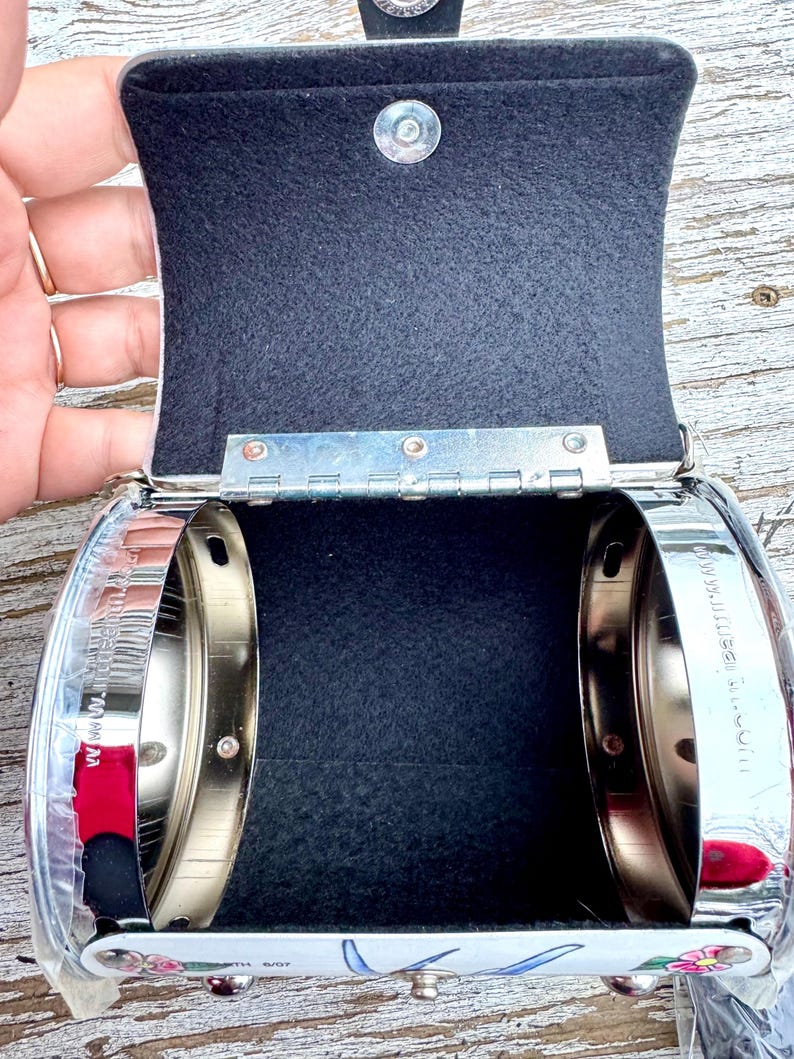 May include: Open, silver-toned metal purse with a black interior. The purse has a hinged lid with a magnetic closure. The bottom of the purse features a floral design with the text "WITH 6/07".