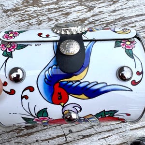 May include: A white, cylindrical container with a decorative tattoo-style design. The design features a blue swallow, pink flowers, and green leaves. The container has a bottle cap closure and a black leather strap.