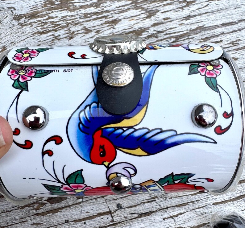May include: A white, cylindrical container with a decorative tattoo-style design. The design features a blue swallow, pink flowers, and green leaves. The container has a bottle cap closure and a black leather strap.