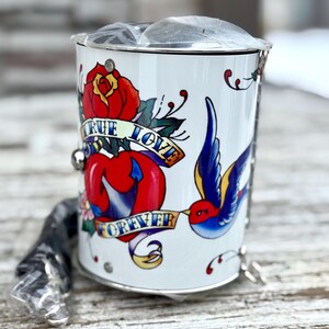 May include: White metal container with a tattoo-style design. The design includes a red rose, a heart, a blue swallow bird, and the words "TRUE LOVE FOREVER" on banners. A silver chain and handle are attached.