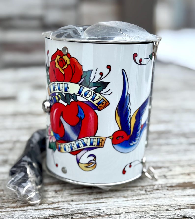 May include: White metal container with a tattoo-style design. The design includes a red rose, a heart, a blue swallow bird, and the words "TRUE LOVE FOREVER" on banners. A silver chain and handle are attached.