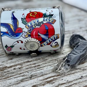 May include: A small, white, cylindrical purse with a tattoo-style design featuring a red rose, a blue swallow, and the words "TRUE LOVE FOREVER." The purse has a silver-colored metal clasp and a black strap.