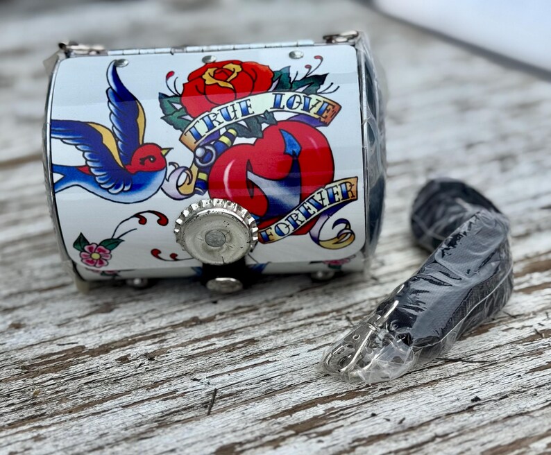 May include: A small, white, cylindrical purse with a tattoo-style design featuring a red rose, a blue swallow, and the words "TRUE LOVE FOREVER." The purse has a silver-colored metal clasp and a black strap.