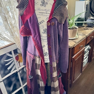 Op de afbeelding: Een lange patchwork cardigan in tinten paars, rood en bruin, tentoongesteld op een mannequin. De trui heeft verschillende texturen en patronen, waaronder strepen en effen kleuren. Het heeft grote houten knopen en een uniek, gelaagd ontwerp, wat een bohemien esthetiek creëert.