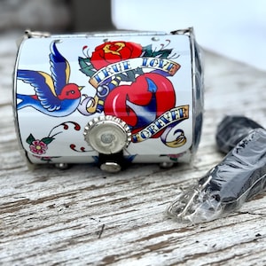 May include: A small, white, metal box with a tattoo-style design featuring a red rose, a blue swallow, and the words "TRUE LOVE FOREVER." The box has a bottle cap closure and a black strap. A black strap is next to the box.