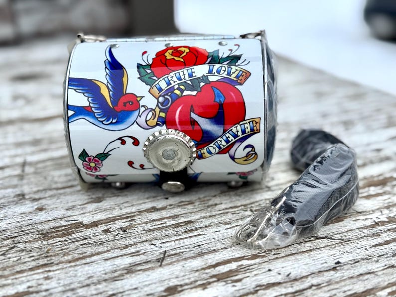 May include: A small, white, metal box with a tattoo-style design featuring a red rose, a blue swallow, and the words "TRUE LOVE FOREVER." The box has a bottle cap closure and a black strap. A black strap is next to the box.