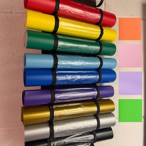 Puede incluir: Una colecci&oacute;n de l&aacute;minas de vinilo enrolladas en varios colores, incluyendo rojo, amarillo, verde, azul, morado, dorado, plateado y negro. Cada rollo est&aacute; asegurado con una correa negra y almacenado en un soporte de pl&aacute;stico transparente. Se ven muestras de color.