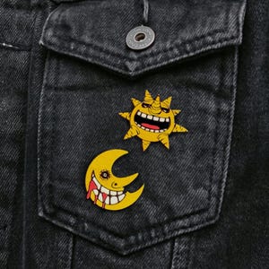 May include: Two yellow enamel pins, one depicting a smiling sun with a horn and the other a smiling crescent moon with blood dripping from its mouth. The pins are on a black denim jacket pocket.