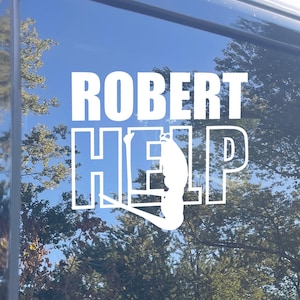 Può includere: Decalcomania bianca su una finestra con le parole "ROBERT HELP" in un carattere grassetto, senza grazie. La parola "HELP" presenta una silhouette stilizzata di una persona. Lo sfondo mostra un cielo blu e alberi.