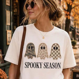 May include: A white short-sleeved t-shirt featuring three ghost illustrations with different patterns: camouflage, leopard print, and a beige checkerboard. Below the ghosts, the text "SPOOKY SEASON" is printed in a bold, black, serif font.