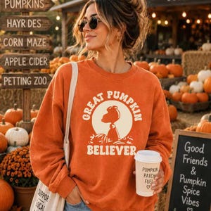 Pode incluir: Uma camisola laranja de manga comprida com um gráfico da silhueta de um cão de desenho animado dentro de um círculo creme com o texto "GREAT PUMPKIN BELIEVER" impresso em laranja. A camisola é usada com um saco de pano branco.