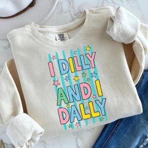 K&ouml;nnte beinhalten: Cremefarbenes Sweatshirt mit dem Text "I Dilly and I Dally" in bunten Blockbuchstaben. Der Text wird durch vertikale blaue Streifen und Sterne erg&auml;nzt. Das Sweatshirt hat einen Rundhalsausschnitt und lange &Auml;rmel.