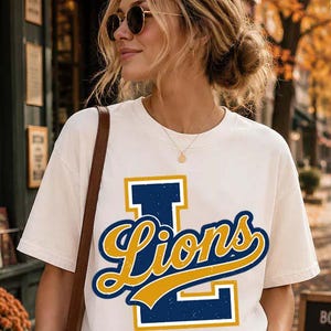 May include: A cream-coloured short-sleeve t-shirt featuring a large navy blue letter L with a distressed texture. The word Lions is printed across the letter in a yellow cursive script with a navy blue outline, creating a bold graphic design.