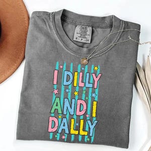 K&ouml;nnte beinhalten: Ein graues T-Shirt mit dem Text "I DILLY AND DALLY" in bunten Buchstaben und Sternen. Das Shirt hat einen Rundhalsausschnitt und ein kleines Etikett am Kragen. Eine silberne Halskette mit Anh&auml;nger liegt &uuml;ber dem Shirt.