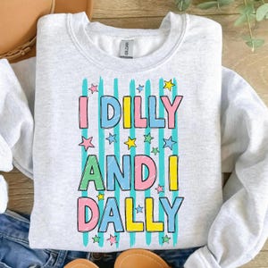 K&ouml;nnte beinhalten: Hellgraues Sweatshirt mit dem Text "I Dilly and I Dally" in bunten Blockbuchstaben. Der Text ist mit Sternen und vertikalen Streifen verziert. Ein l&auml;ssiges, bequemes Oberteil.