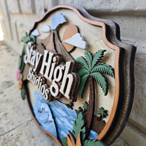 May include: A multi-layered wooden sign featuring the text Stay High Studios in white lettering. The design includes a brown volcano, a yellow sun, white clouds, green palm trees, and blue ocean waves, all set against a light wood background.