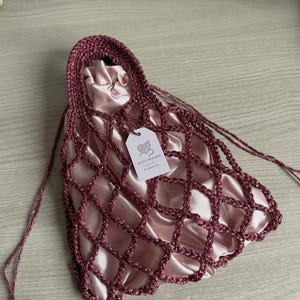 Può includere: Una borsa a rete all'uncinetto rosa scuro con una trama di filato metallizzato scintillante e un manico solido. All'interno della struttura a rete si trova una pochette in raso rosa chiaro con coulisse. Un'etichetta bianca riporta il testo ALMA CROCHET.