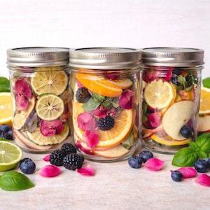 May include: Three glass mason jars filled with dried citrus slices, rose petals, and berries. The jars are sealed with silver lids and sit on a light-colored surface, surrounded by fresh basil and scattered berries.