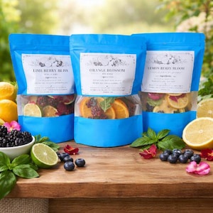 May include: Three blue pouches with clear windows, each labeled with a different fruit blend: Lime Berry Bliss, Orange Blossom, and Lemon Berry Bloom. The pouches are filled with dried fruit slices and berries. Fresh fruit and flowers are arranged around the pouches on a wooden surface.